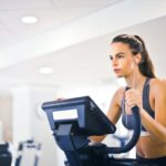 Serious fit woman in earphones and activewear listening to music and running on treadmill in light contemporary sports center