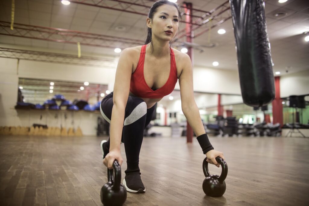 From below focused young Asian female athlete in sportswear squatting with kettlebells while training alone against blurred interior of light modern gym