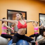 Serious slim ethnic female athlete in sportswear sitting on fit ball with arms outstretched and stretching body while training with other ladies in contemporary fitness center